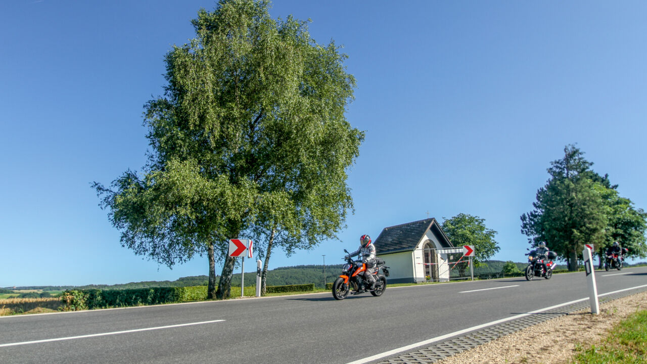 Motorräder auf einer Landstraße mit Kurvenpfeil, daneben ein kleines weißes Haus und eine Birke
