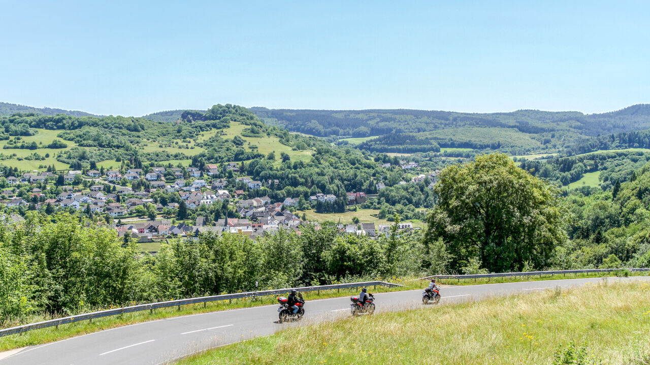 Drei Motorräder auf einer Straße am Hang, darunter ein Ort im Tal zwischen bewaldeten Kuppen
